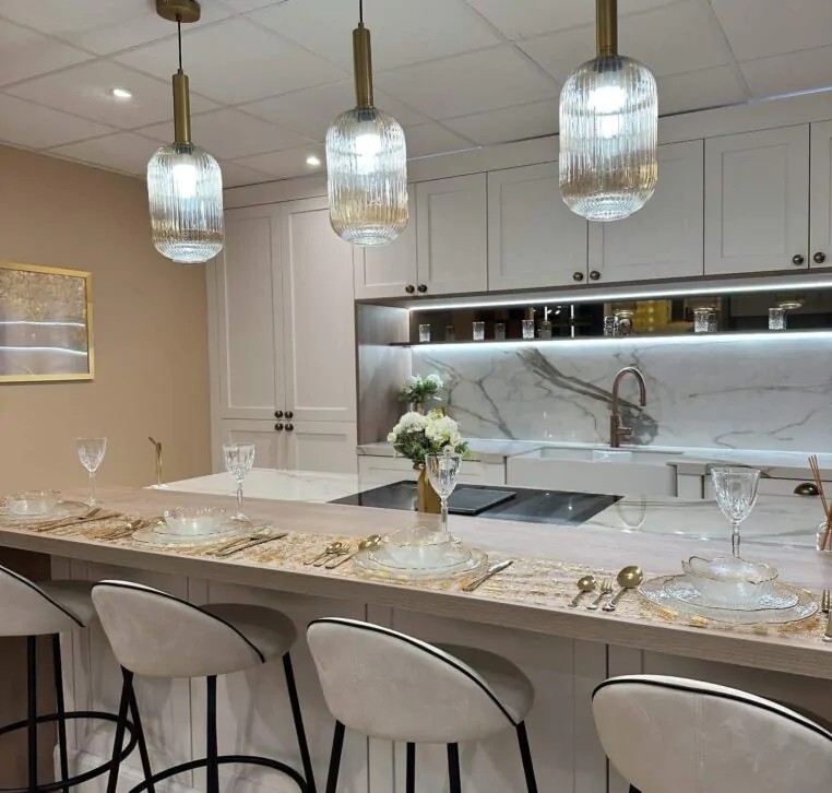 Elegant white kitchen with gold accent lighting, marble island, and chic table setting.