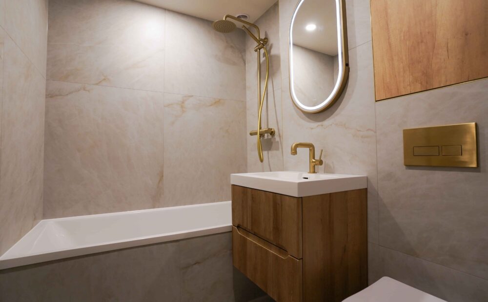 Completed bathroom renovation showing the bathtub, fluted wood vanity, and brushed gold shower set.