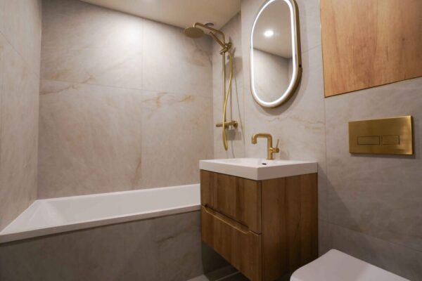 Completed bathroom renovation showing the bathtub, fluted wood vanity, and brushed gold shower set.