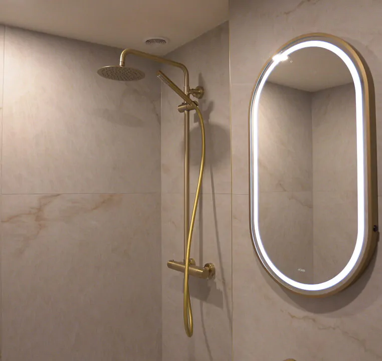 Bathroom view showing the coordination between the brushed gold shower system and the oval LED mirror.