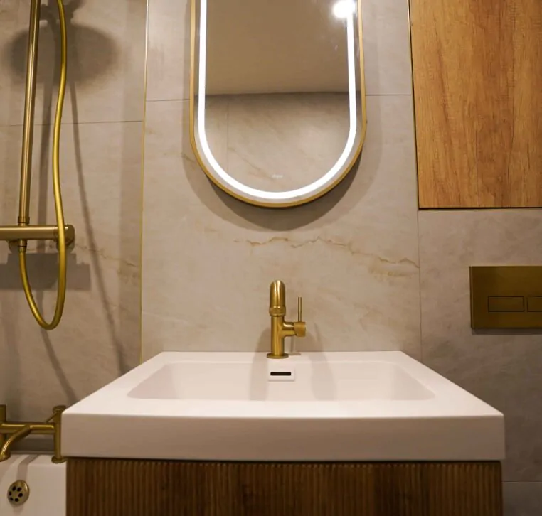 Oval LED backlit bathroom mirror with a brushed gold frame mounted on a neutral stone-effect tiled wall.
