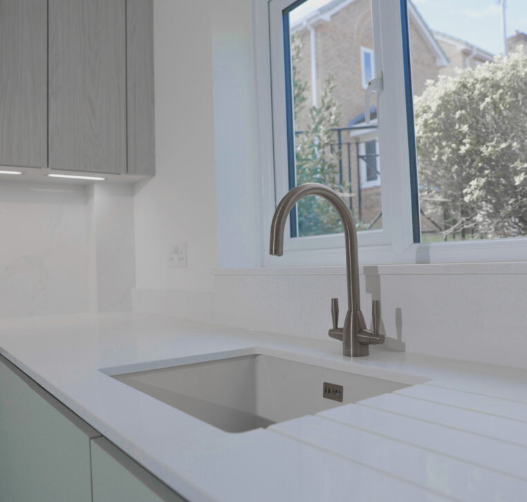 Close-up of modern kitchen sink with brushed metal mixer tap and white worktop beneath window