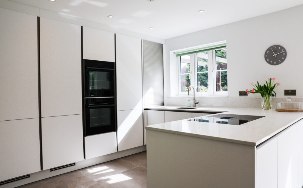 White handleless kitchen with large island-style worktop, integrated hob, built-in ovens and garden views
