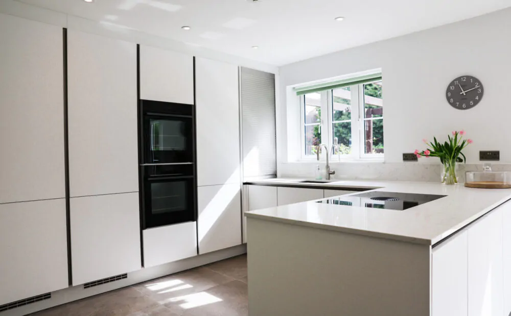 White handleless kitchen with large island-style worktop, integrated hob, built-in ovens and garden views